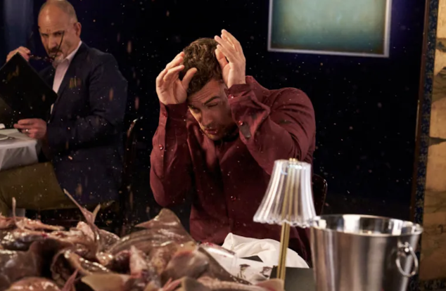 Theo James shields himself from flying food at a fancy dining table covered with fish, amidst a chaotic scene.