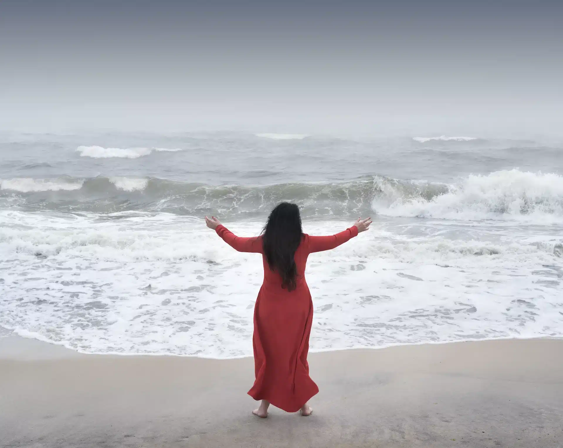 A woman in a flowing red dress stands barefoot at the shoreline, arms outstretched towards the misty sea and fading horizon.