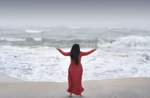 A woman in a flowing red dress stands barefoot at the shoreline, arms outstretched towards the misty sea and fading horizon.