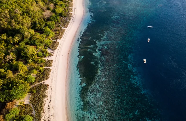 Aerial view of a sandy beach bordered by lush greenery and clear turquoise waters, with small boats anchored offshore.